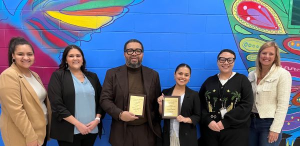 Six people standing in front of a butterfly mural, the center two are holding award plaques