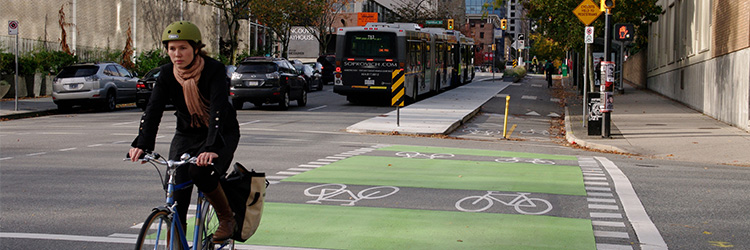 A woman is cycling down a city street with buses in the background.